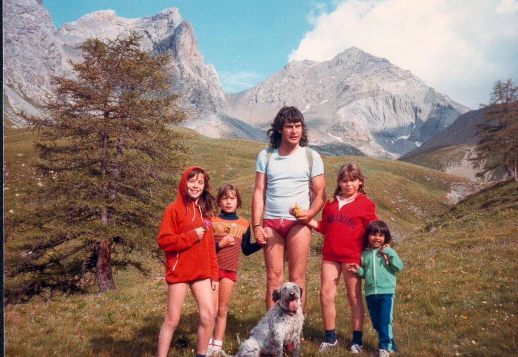 1970s holiday photograph showing a high-mountain landscape with four children, a man, and a dog posing together for the camera
